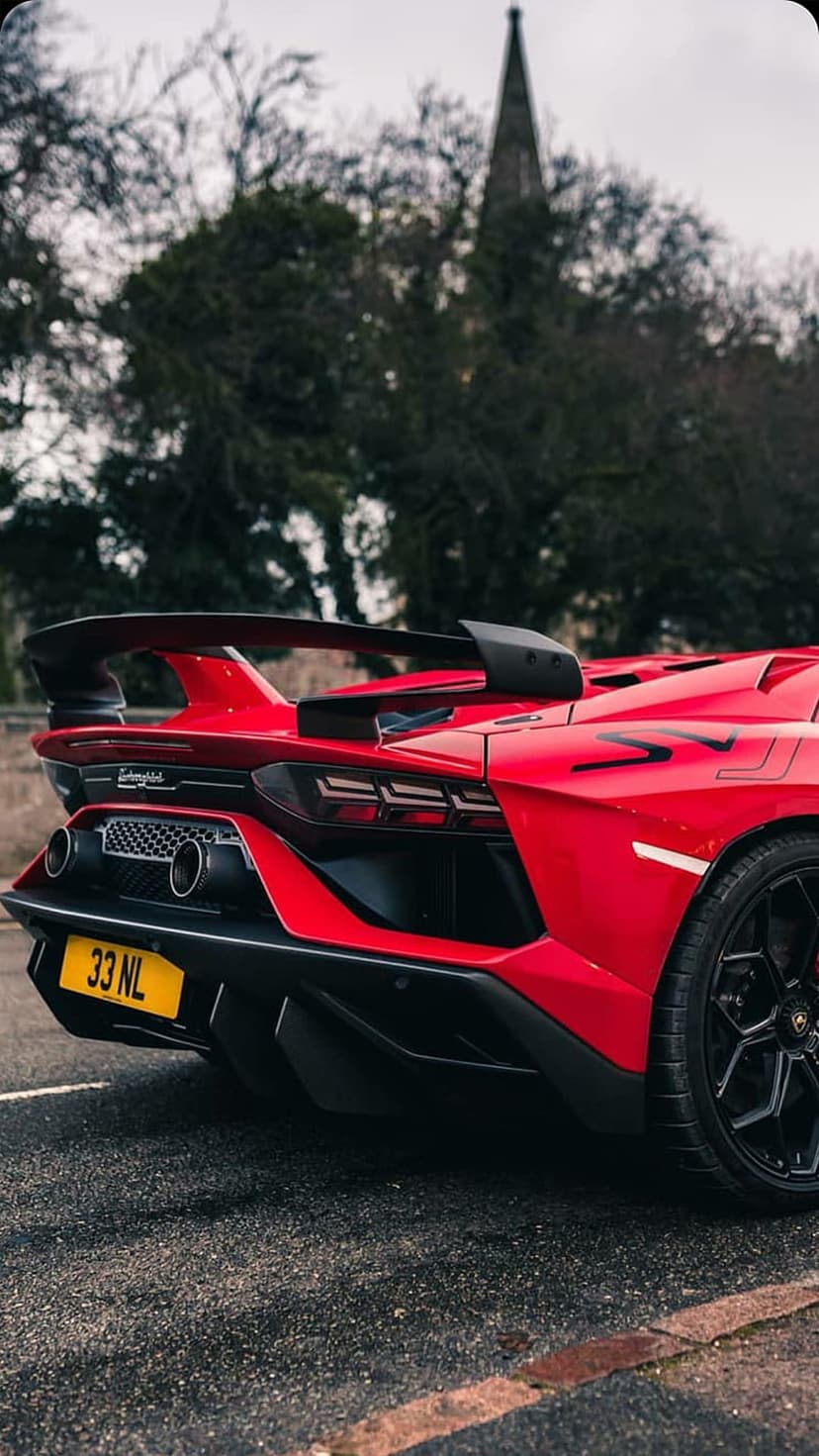 Red Lamborghini SVJ rear wing and taillights on a wet road with a gothic church steeple under a grey sky.