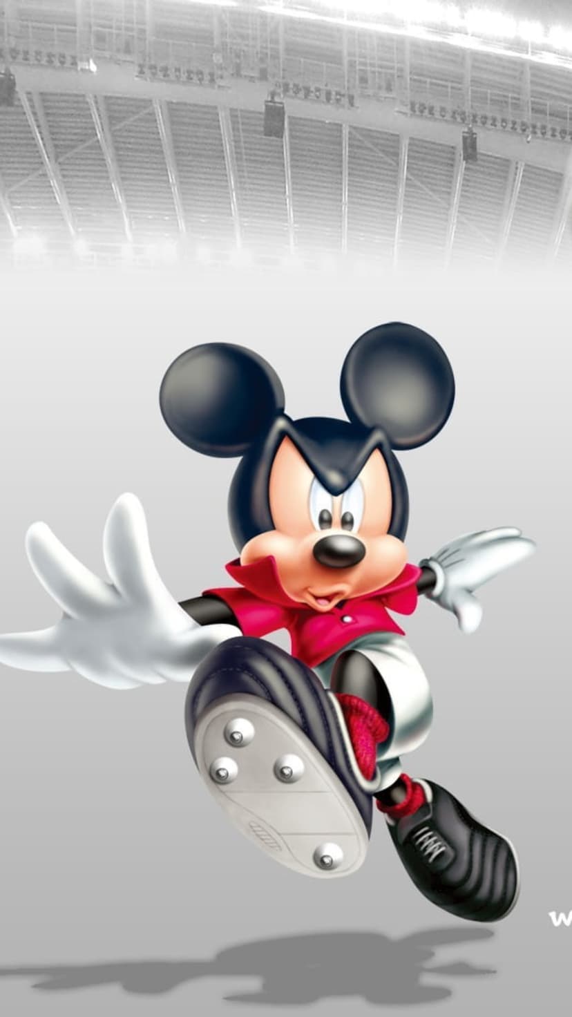 Mickey Mouse wearing a soccer uniform and cleats runs through a blurred monochrome stadium arena in mid-action.