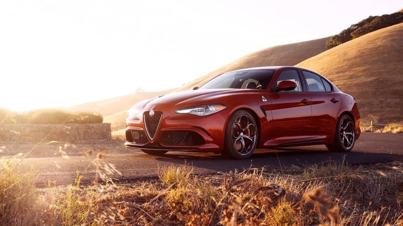 Red Alfa Romeo Giulia Quadrifoglio parked on a winding road with rolling hills and golden hour sunlight flare.