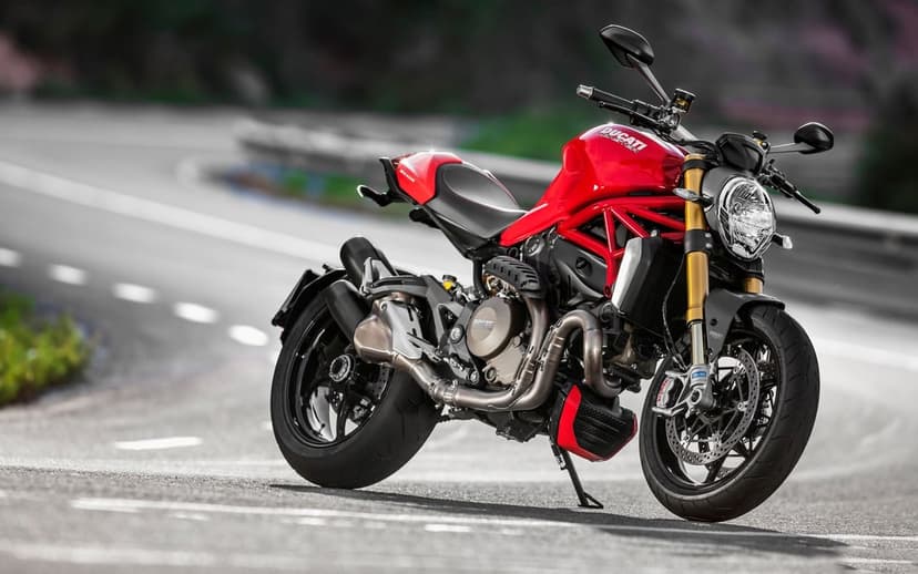 Red Ducati Monster motorcycle parked on a winding asphalt road with a blurred mountain landscape background.