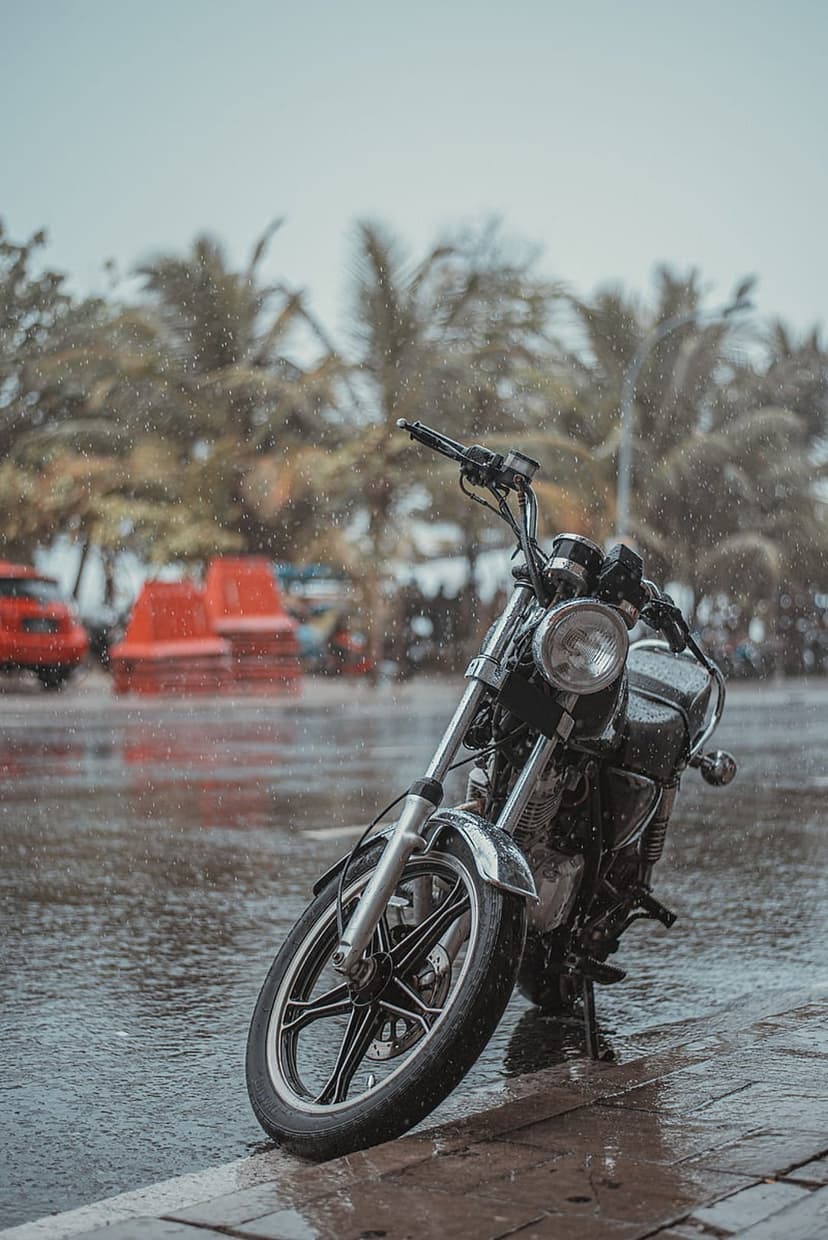 Vintage motorcycle parked on a wet street during heavy tropical rain with palm trees blurred in the background.