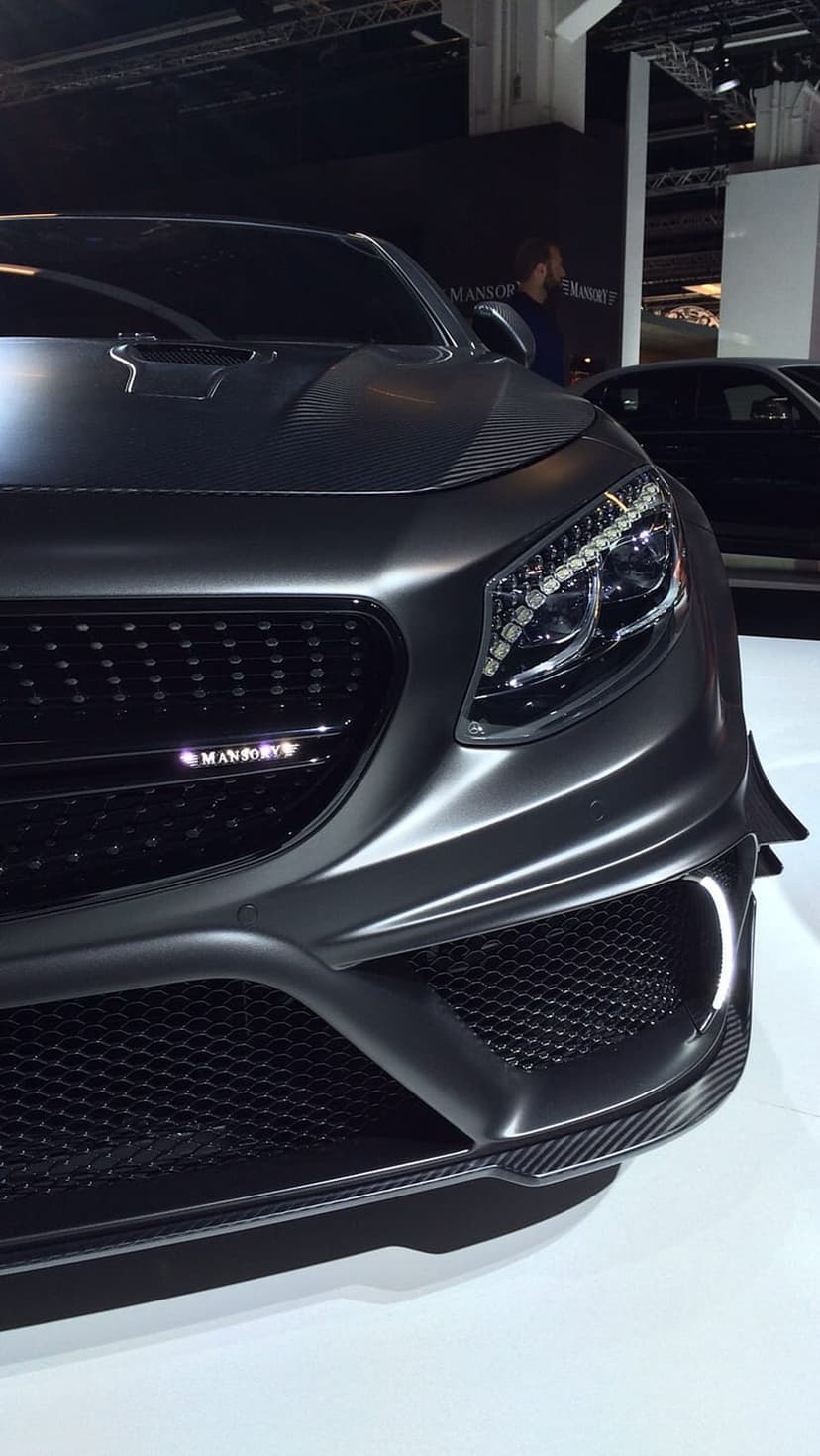 Mansory Mercedes S-Class front grille with carbon fiber accents, LED headlights, and matte grey paint at a car show.