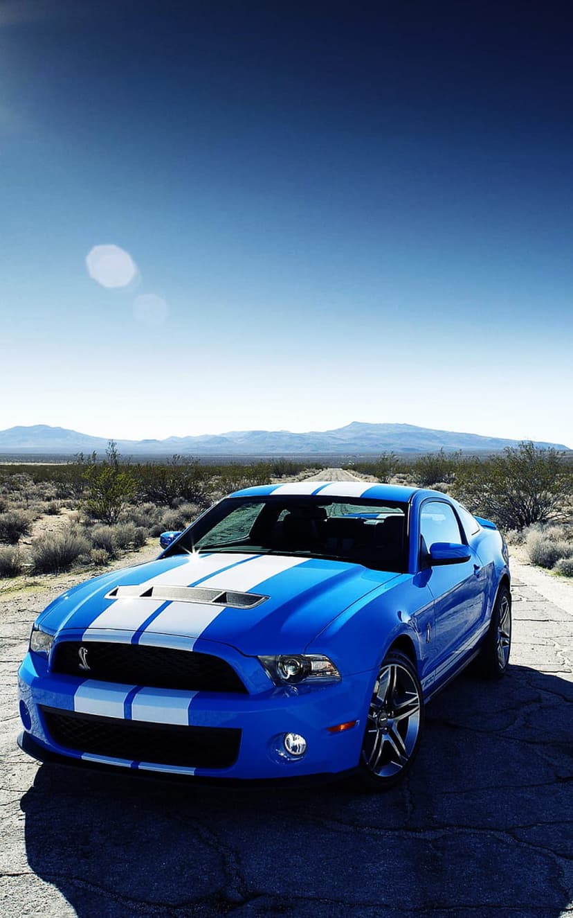 Blue Ford Mustang Shelby GT500 with white racing stripes on a cracked desert road under a bright clear sky.