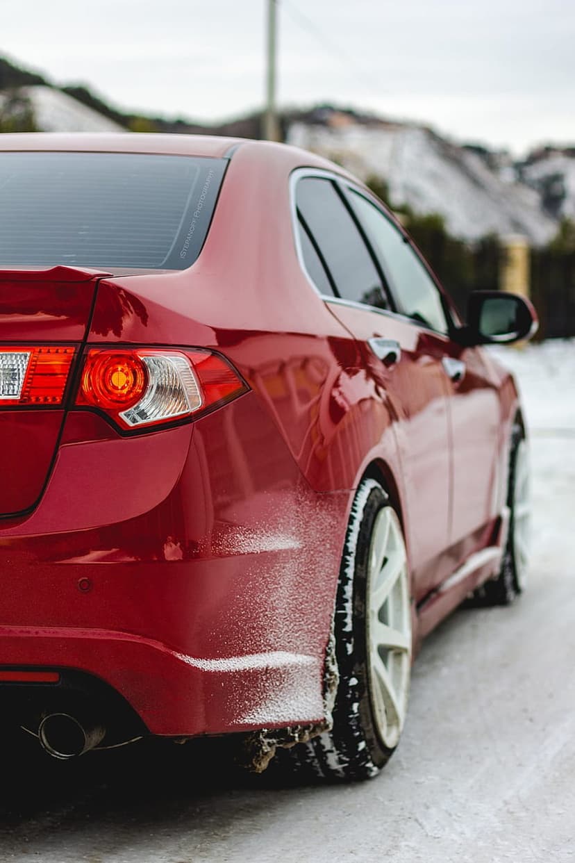 Red sedan rear panel with white wheels parked on a snowy ground against a blurred winter landscape background.