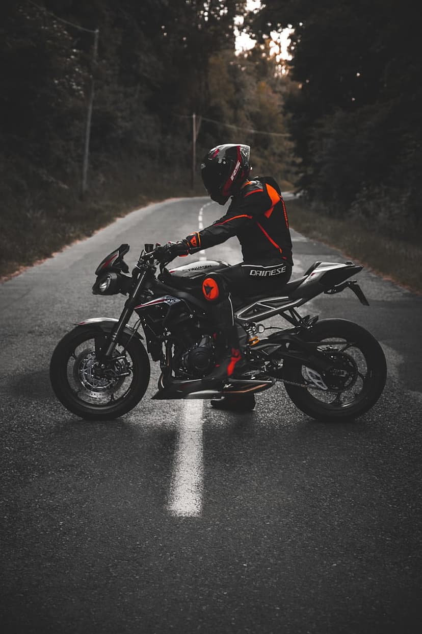 Motorcyclist in black and orange Dainese gear sits on a sportbike atop a wet, sun-dappled road lined with trees.