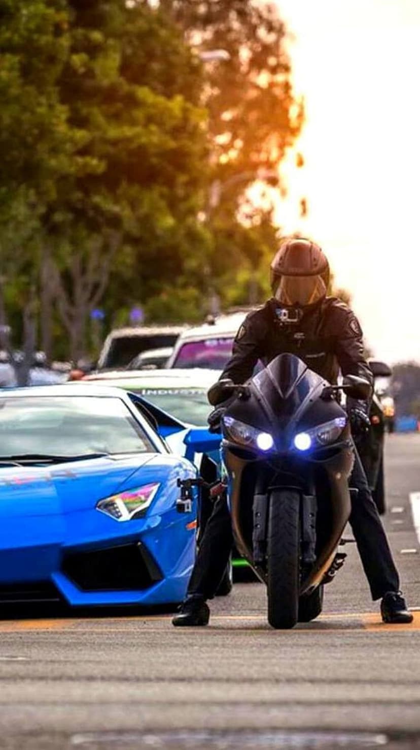 Biker in leather gear stands beside a sapphire blue supercar on an asphalt road under a bright summer sky.