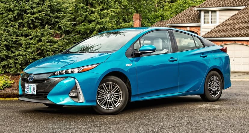 Teal blue Toyota Prius Prime parked on a driveway with green trees and a suburban house in the background.