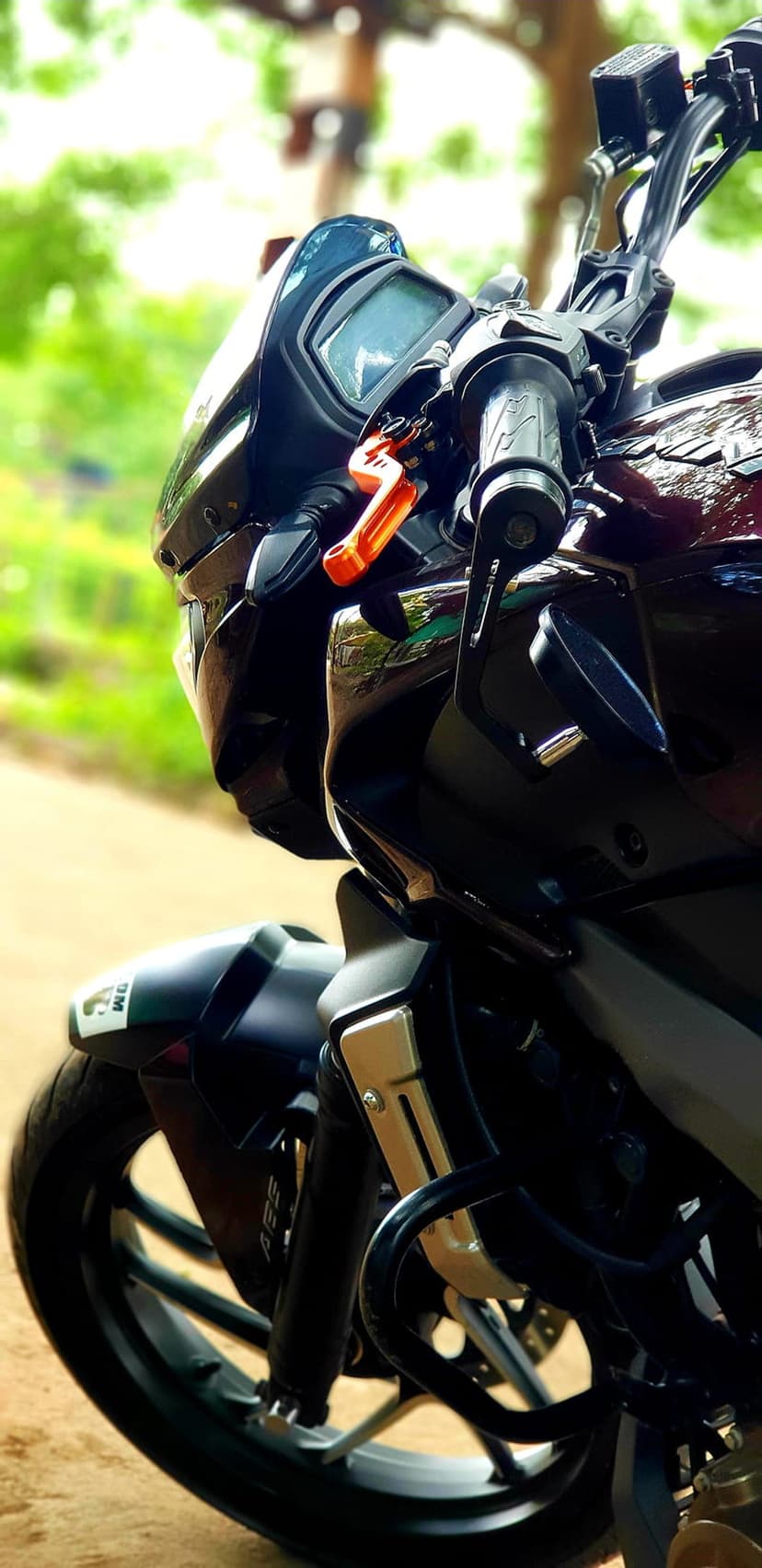 Maroon and black motorcycle front with a digital display, orange brake lever, and ABS wheel against blurry trees.
