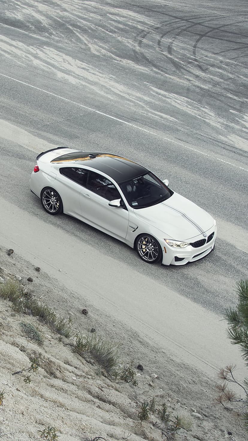 White BMW M4 coupe with racing stripes and a black roof parked on a dusty road with visible tire tracks.
