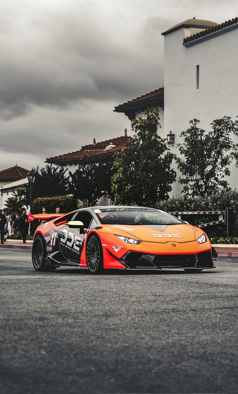 Orange Lamborghini race car with aerodynamic kit parked on asphalt road in front of Spanish style buildings.