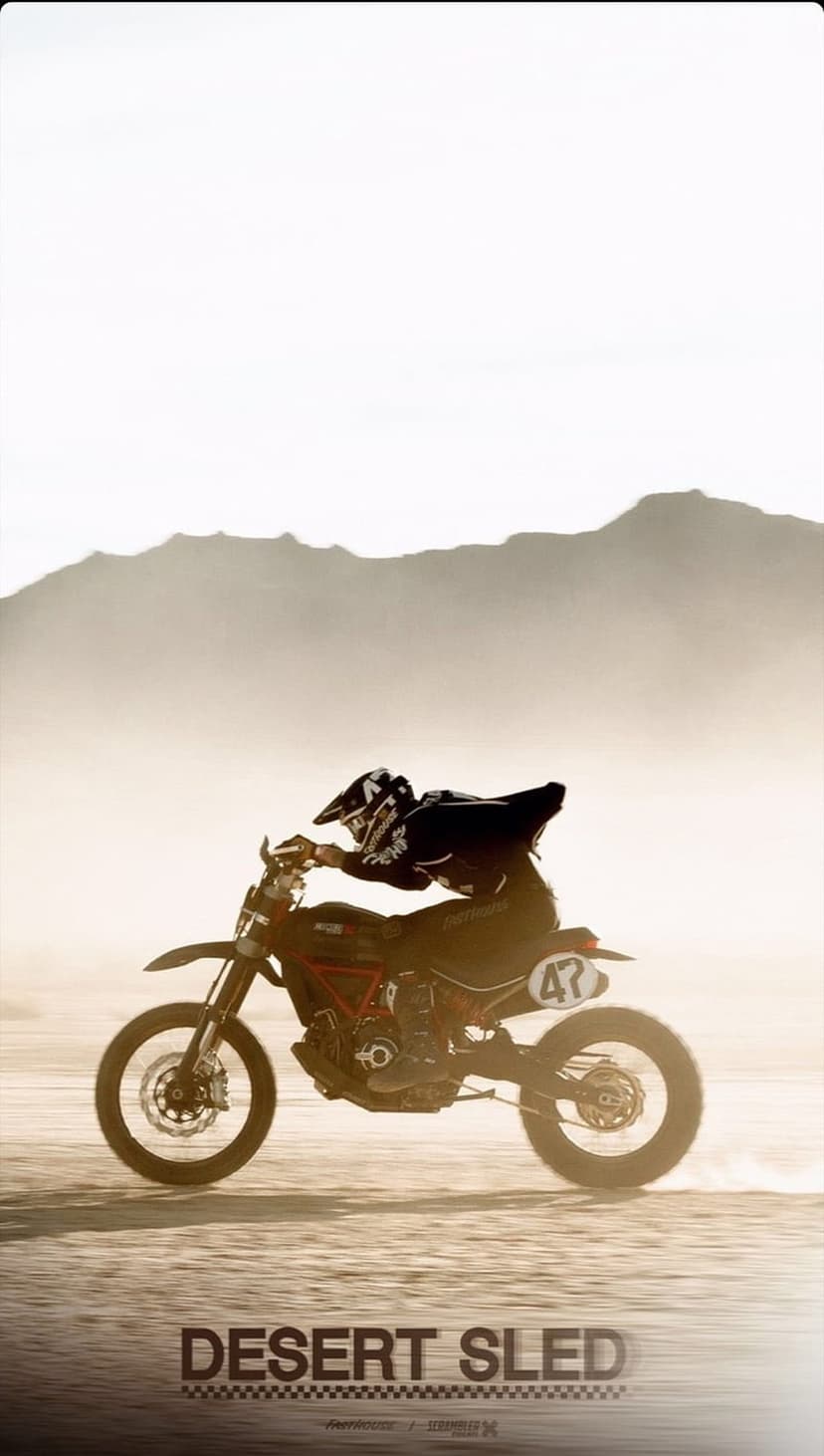 Motocross rider in full gear soaring mid-air over a dusty desert floor with hazy mountains in the background.