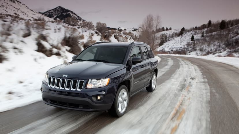 Dark navy Jeep Compass SUV driving on a snow-lined mountain road with blurred scenery and a muted sky.