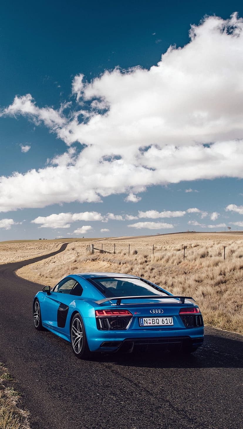 Blue Audi R8 sports car driving on a curved asphalt road through dry grassy fields under a bright cloudy sky.
