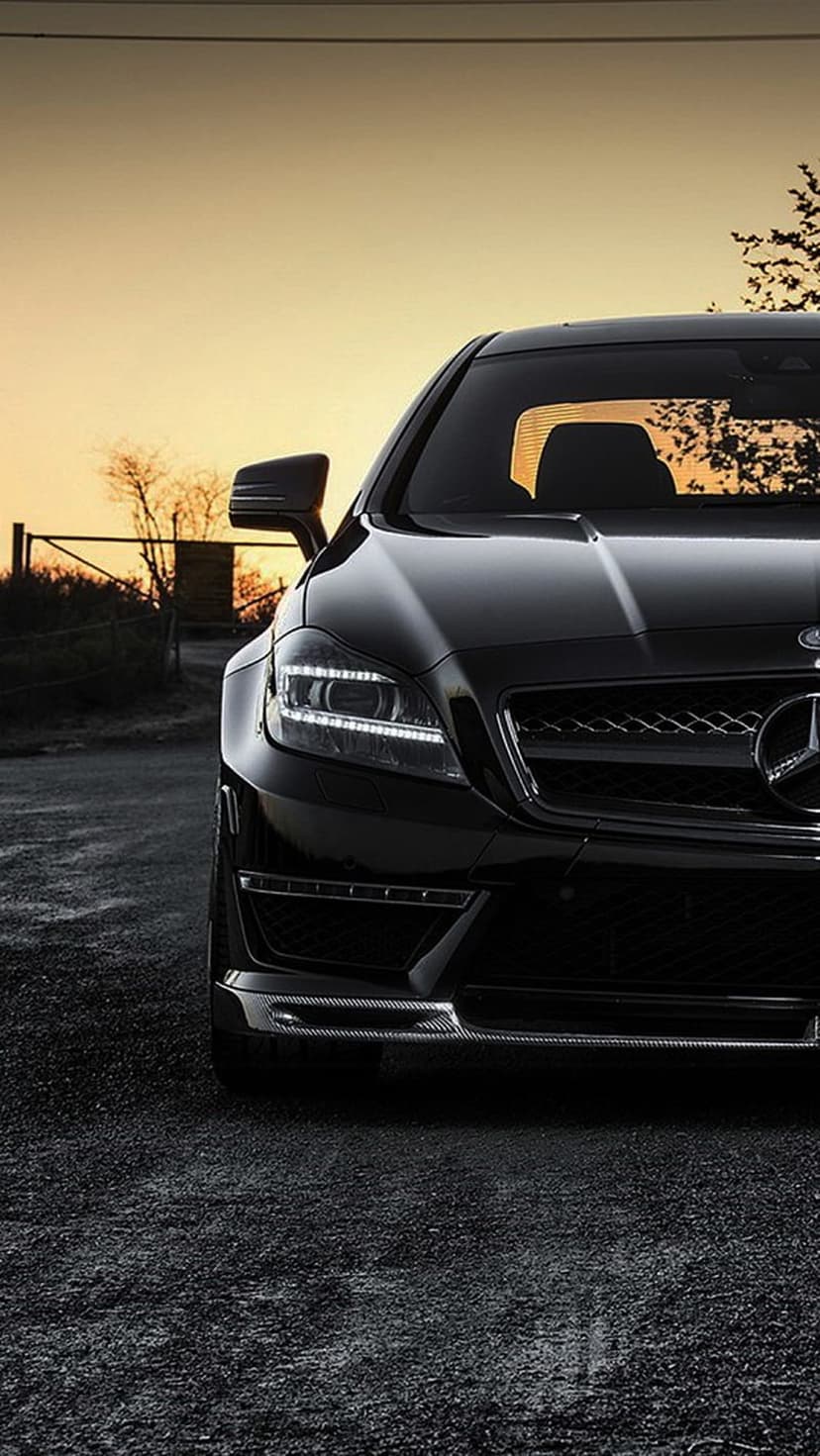 Black Mercedes-Benz CLS AMG with lit LED headlights parked on a gravel road during a vibrant golden sunset.