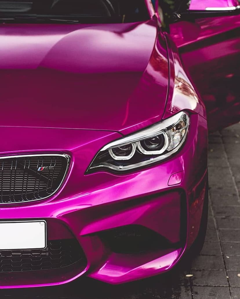 Pink BMW M2 sports car with an open door parked on a textured cobblestone street at a low camera angle.