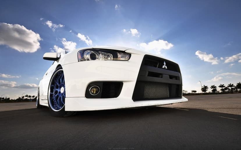White Mitsubishi Lancer Evolution X with blue multi-spoke wheels parked on asphalt under a bright blue sky.