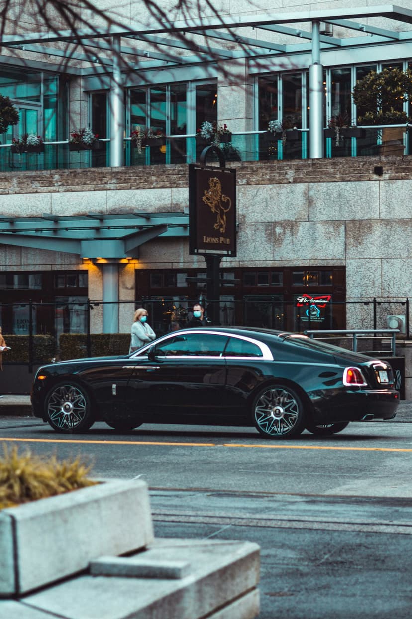Black Rolls-Royce coupe drives past a modern glass building and a pub sign on a bright metropolitan street.