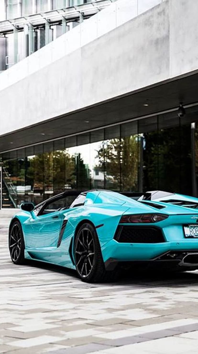 Bright blue Lamborghini Aventador Roadster parked on a patterned stone walkway in front of a glass building.