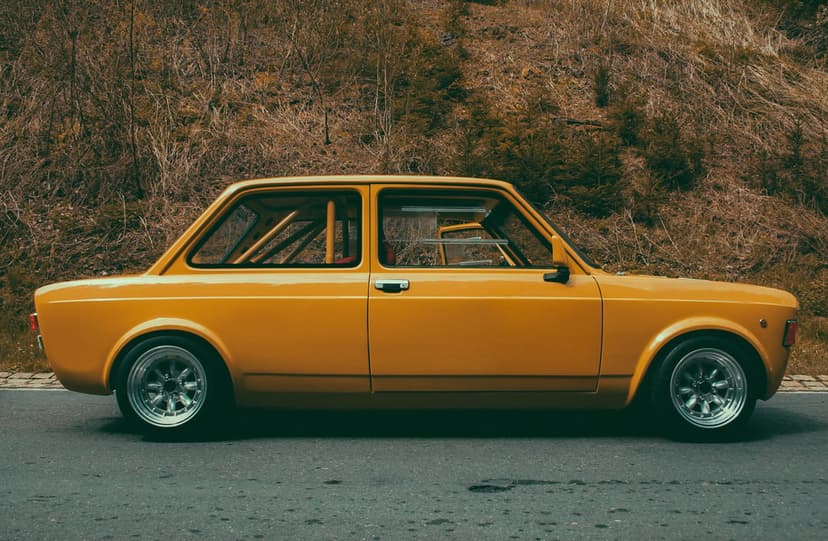 Yellow Fiat 128 Rally car with a roll cage parked on an asphalt road against a dry textured hillside backdrop.
