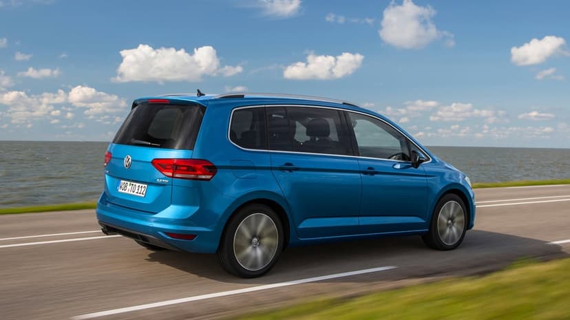 Blue Volkswagen Touran driving on a paved road beside a calm sea under a bright blue sky with white clouds.