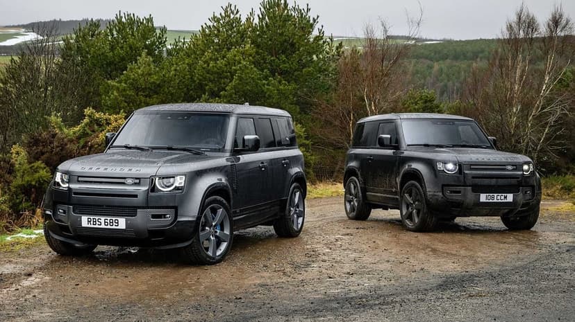 Two grey Land Rover Defenders parked on a muddy path in a rainy forest with pine trees and rolling green hills.