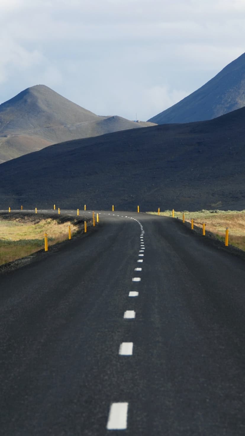 Asphalt road with yellow markers stretches toward dark volcanic mountains under a bright overcast sky.