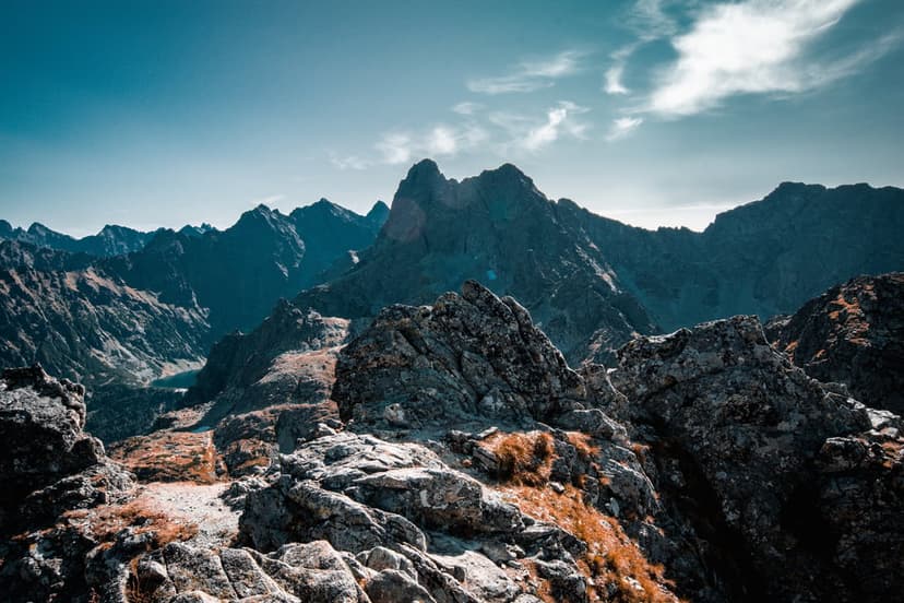 Sharp gray mountain peaks rise above dry golden grass and weathered boulders under a clear blue sky with thin clouds.