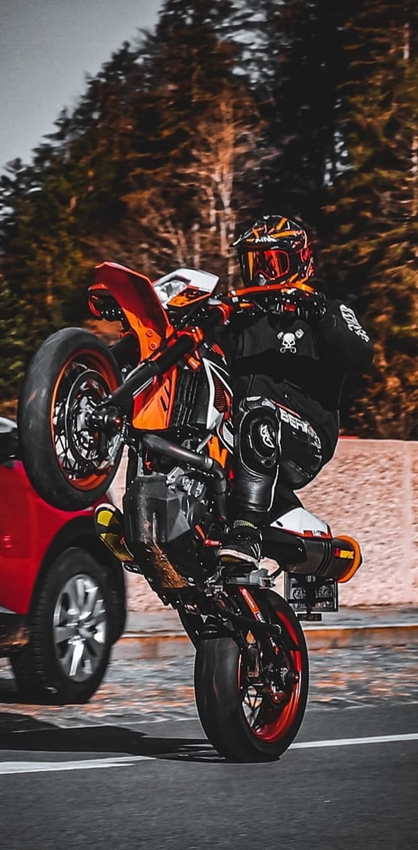 Motorcyclist in black gear performs a wheelie on an orange bike beside a blurred red car on a paved road.