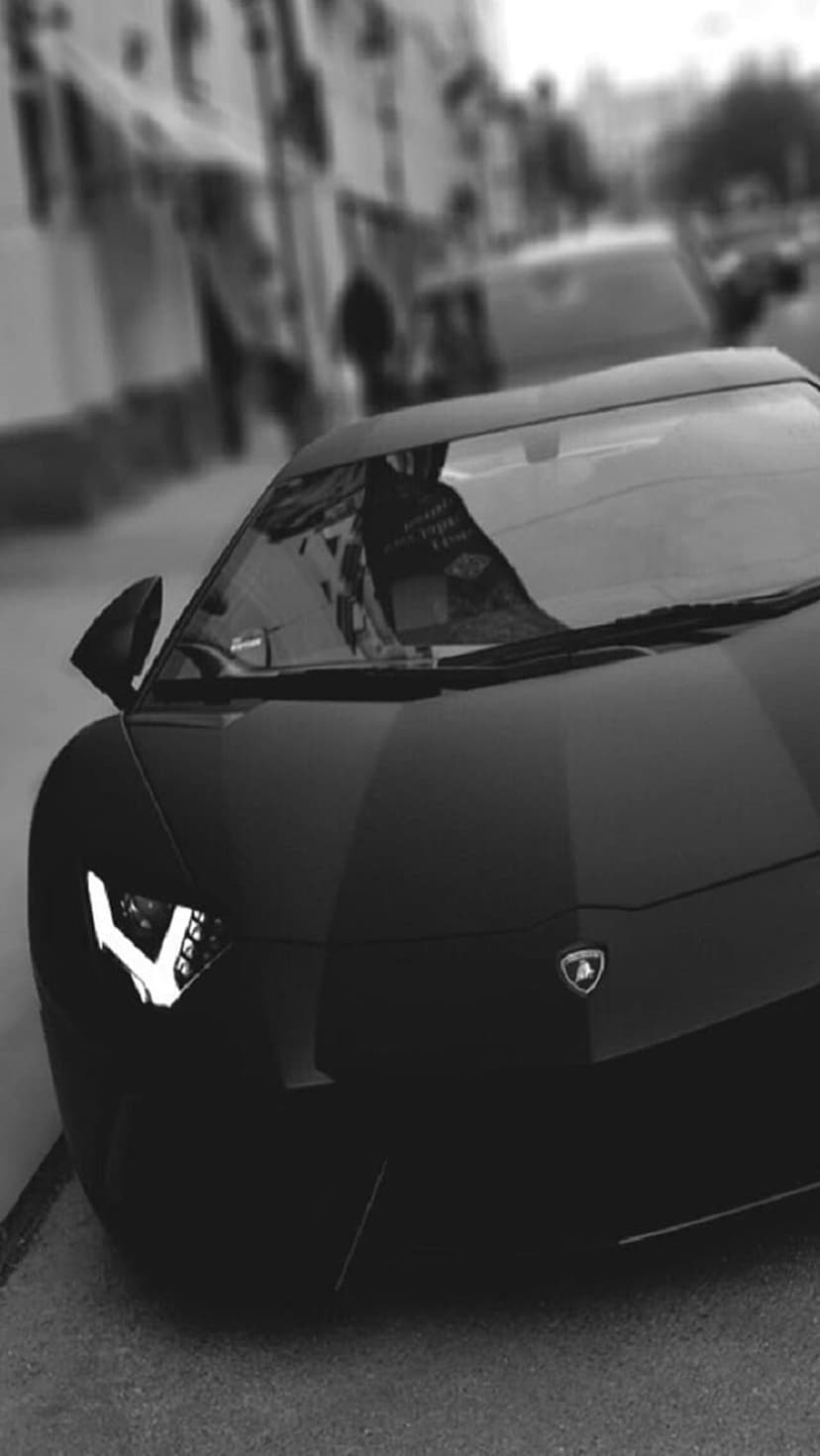 Matte black Lamborghini Aventador with illuminated Y-shaped headlights parked on a blurred urban street at night.