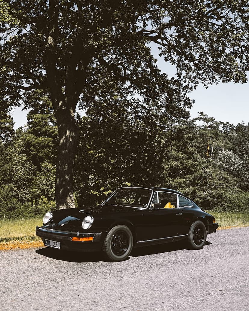 Black Porsche 911 with round headlights parked on a gravel road under a massive oak tree with green leaves.