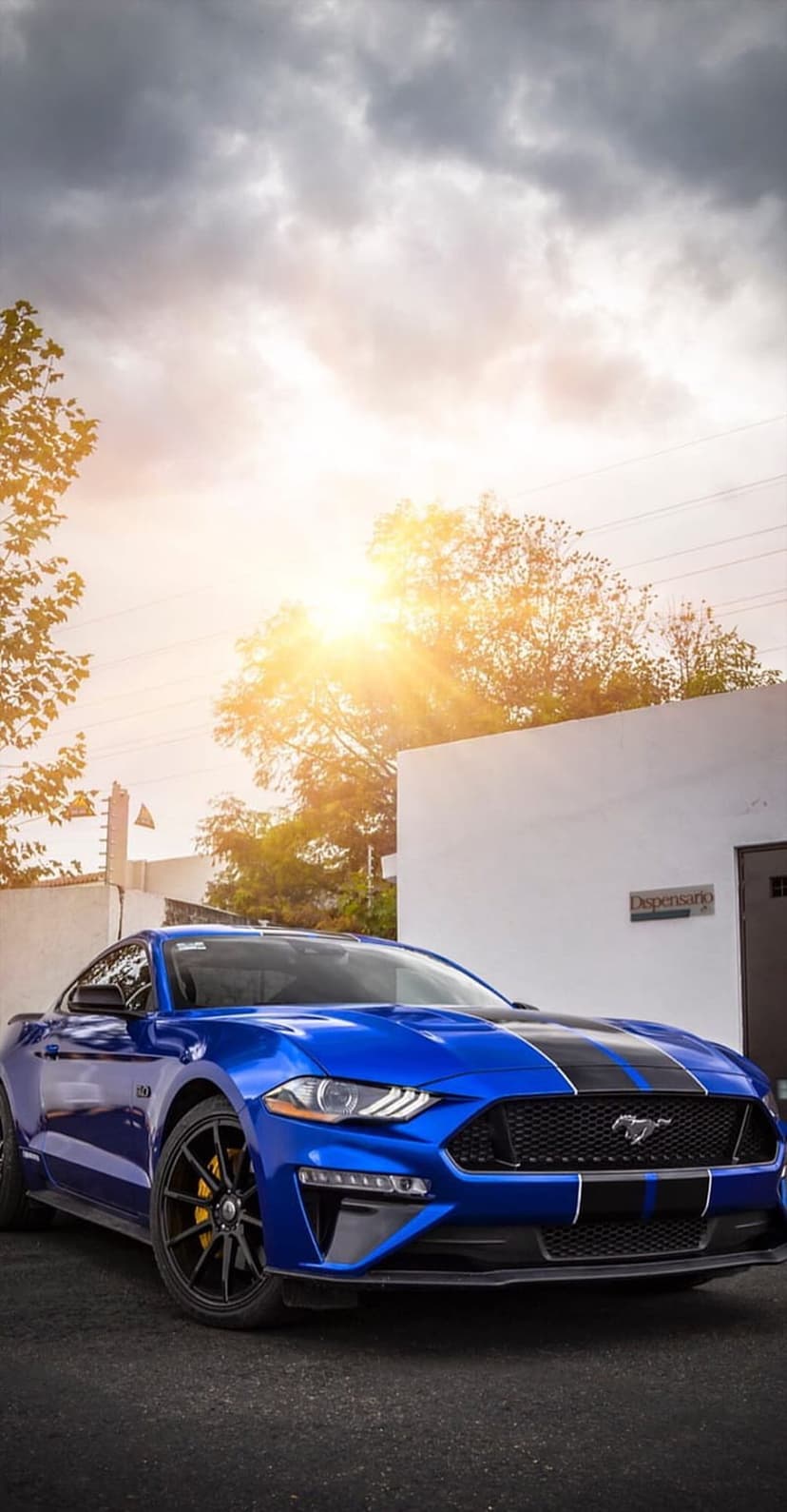 Blue Ford Mustang GT with black racing stripes parked on asphalt outdoors under a sunlit cloudy sky.