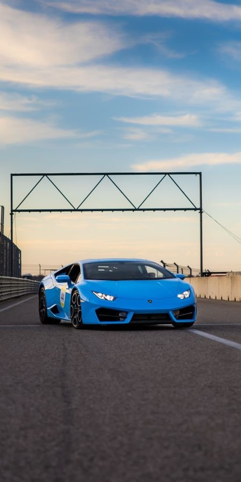 Blue Lamborghini Huracan racing on asphalt under a sunset sky beneath a large triangular metal structure.