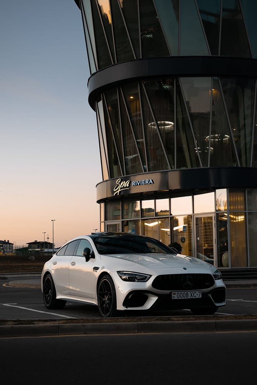 White Mercedes-AMG GT 4-door coupe with black wheels parked near a curved glass building during twilight.