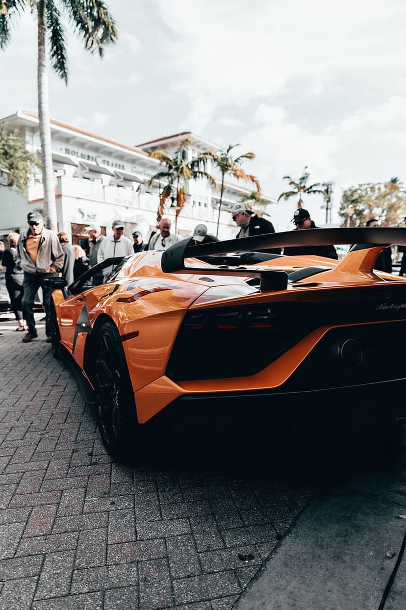 Orange Lamborghini Aventador SVJ with a large rear wing parked on a brick road under coastal palm trees.