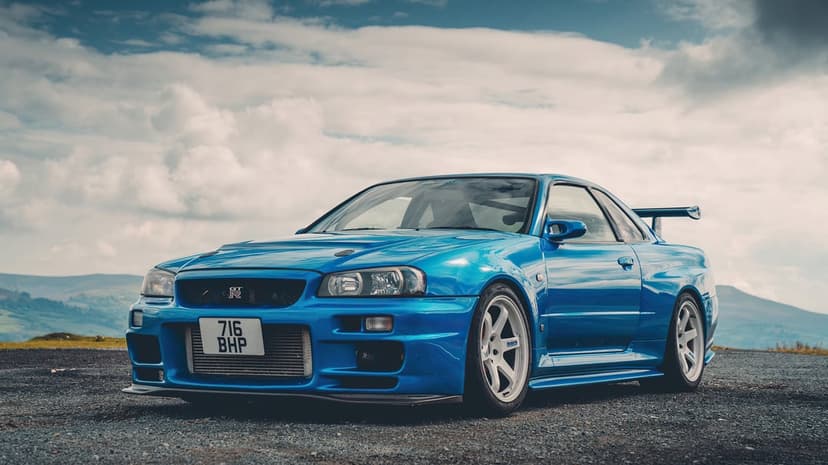 Blue Nissan Skyline GT-R R34 with white wheels parked on gravel under a cloudy sky in the mountains.