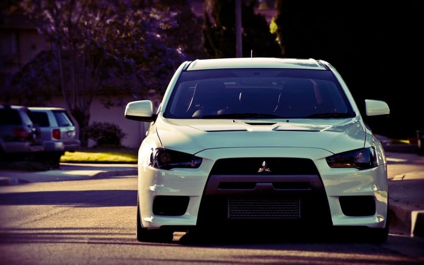 White Mitsubishi Lancer Evolution X front view parked on an asphalt street with purple trees at sunset.