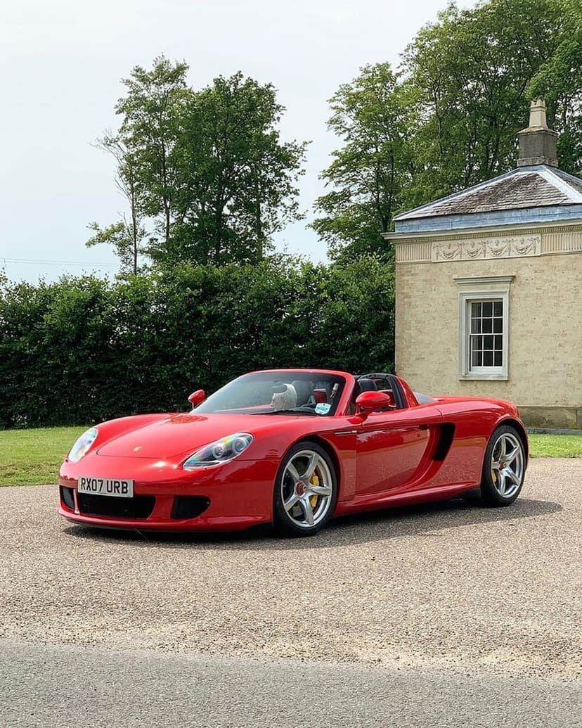 Red Porsche Carrera GT convertible with yellow calipers parked on a gravel driveway against a leafy green backdrop.