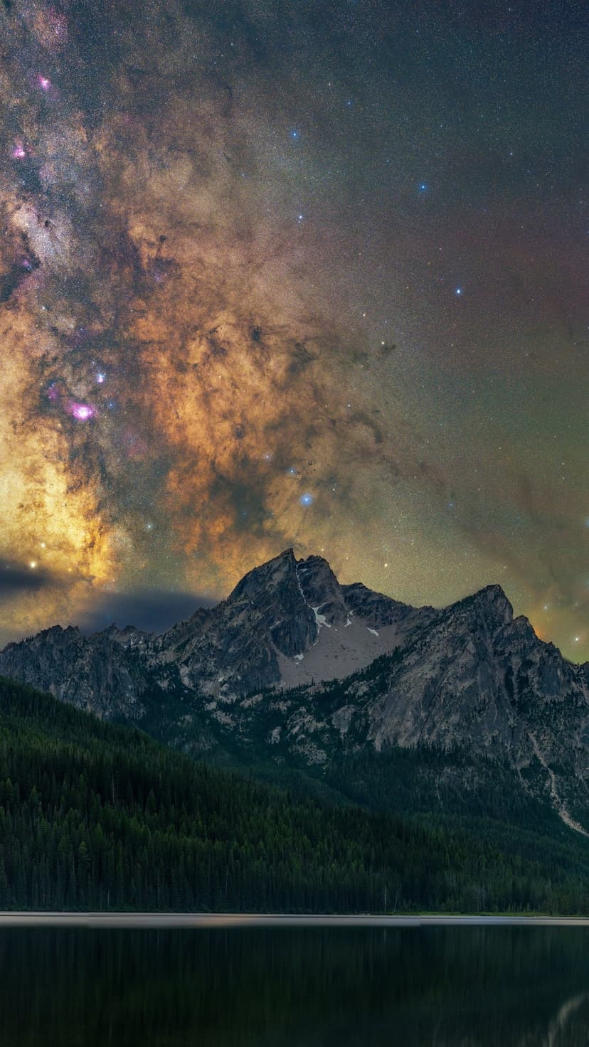 Luminous Milky Way galaxy arches over a jagged mountain peak and still alpine lake at night in the Idaho backcountry.