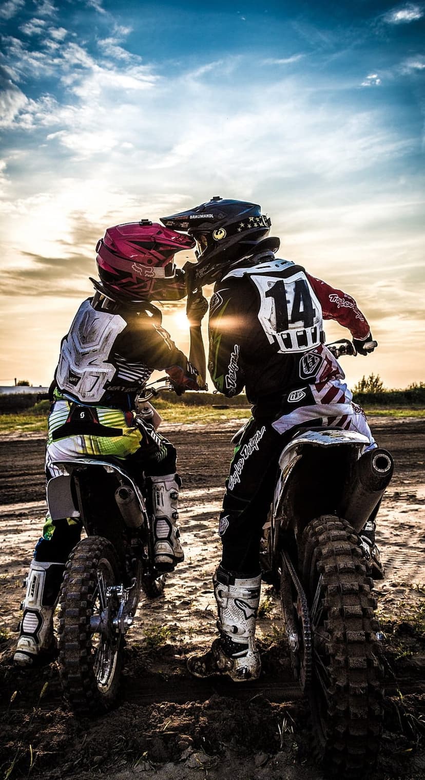 Motocross riders in racing gear kiss on motorcycles beneath a radiant golden hour sunset on a dirt track.