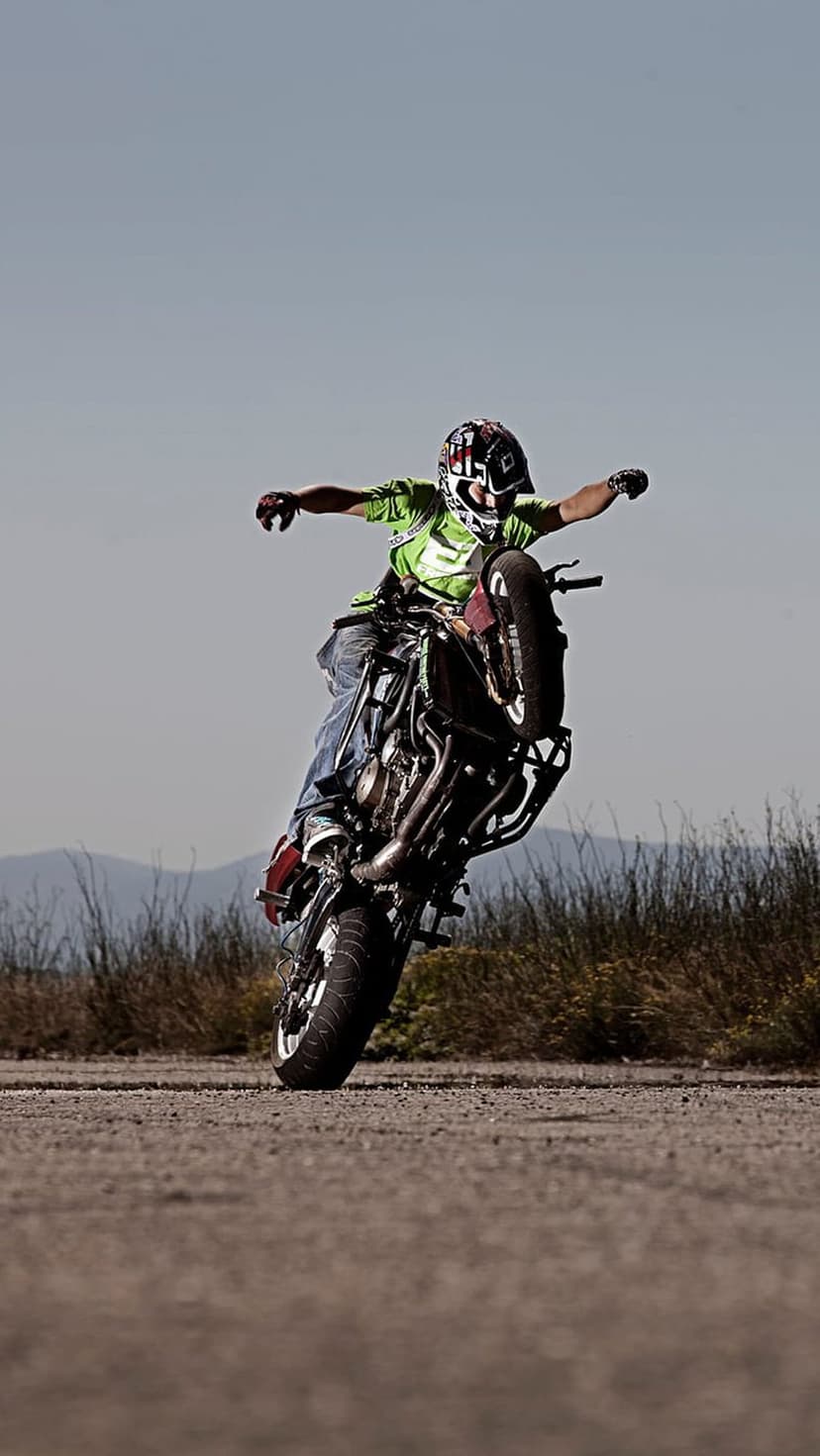 Motorcyclist in a green jersey performs a wheelie on a dirt path with mountains under a clear blue sky.