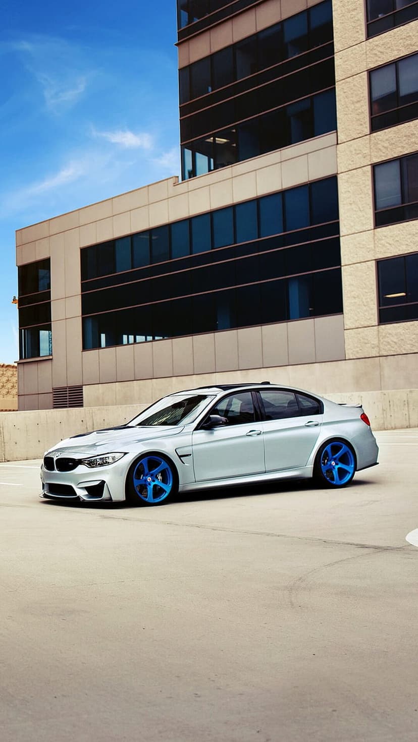 Silver BMW M3 sedan with electric blue wheels parked on concrete beside a modern glass and steel building.