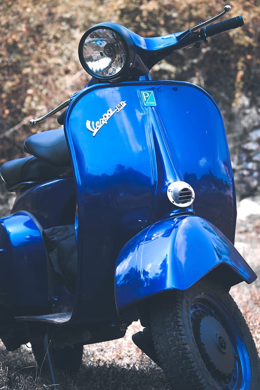 Blue Vespa 150 scooter with chrome headlight and glossy paint parked on dry grass during a bright day.