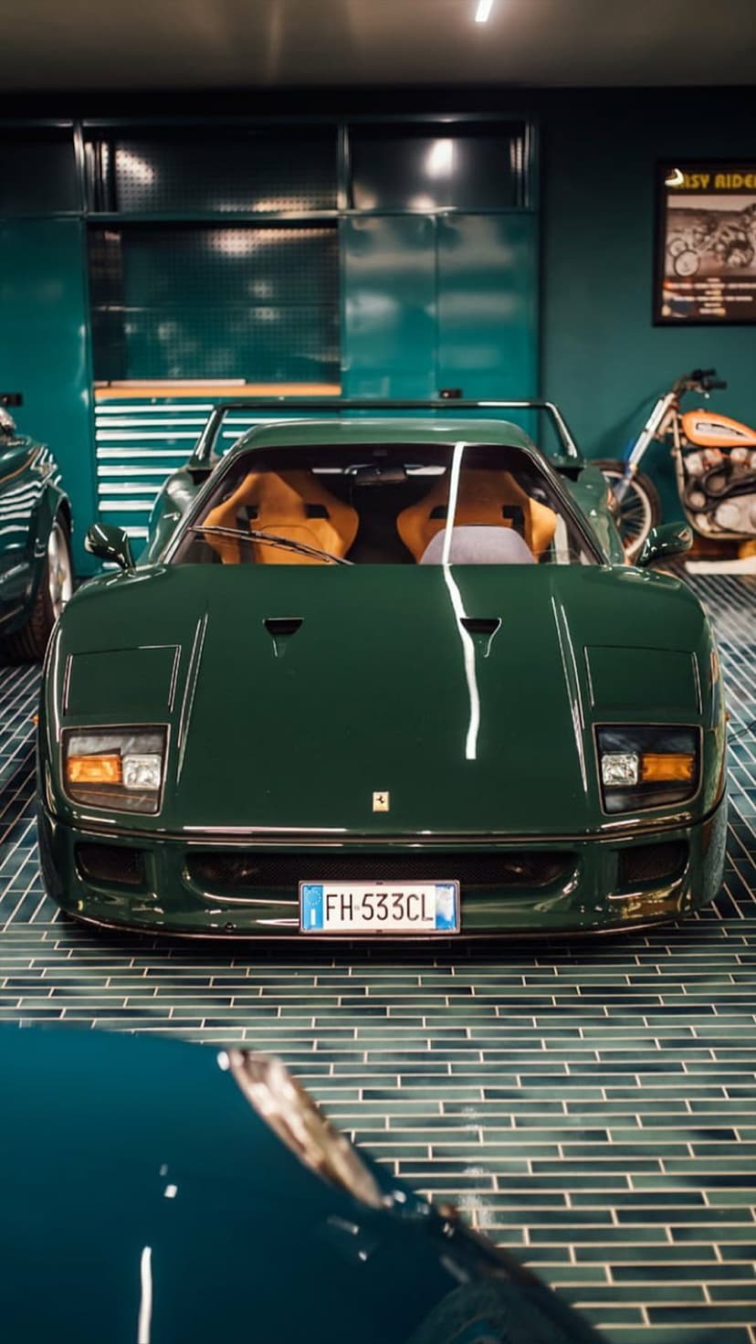 Dark green Ferrari F40 with tan leather seats parked in a modern garage near a vintage motorcycle and teal cabinets.