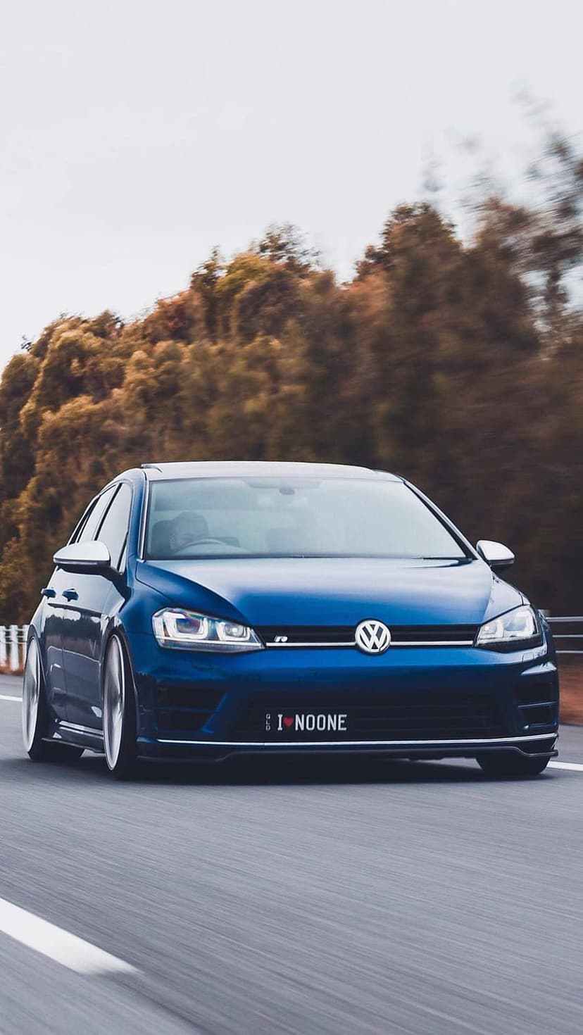 Blue Volkswagen Golf R driving on a highway with motion-blurred autumn trees and a custom I NOONE license plate