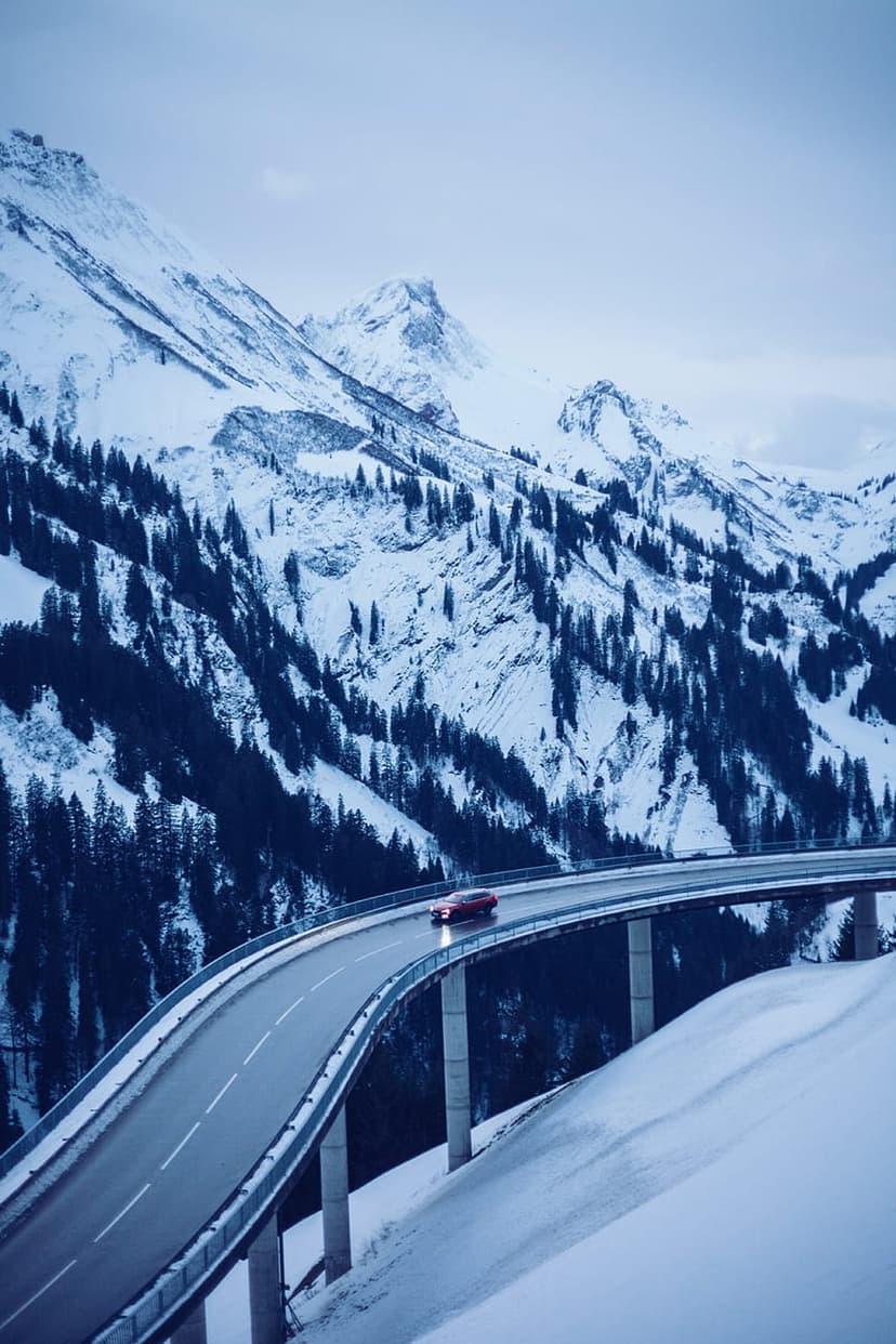 Red car driving on a winding road through snow-covered mountains and evergreen forests in winter.