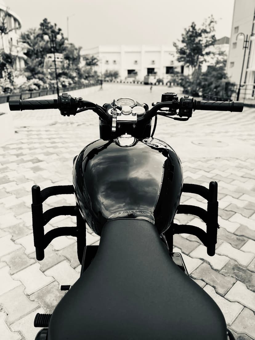 Royal Enfield motorcycle front view showing handlebars, fuel tank, and engine guard in a monochromatic setting.