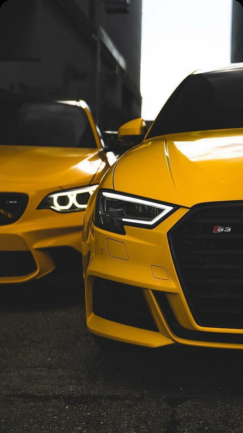Two yellow Audi RS3 cars feature sharp LED headlights and honeycomb grilles against a dark, shadowy background.