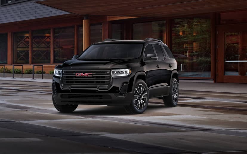 Black GMC Acadia SUV parked in front of a modern wood and glass building on a damp urban pavement at twilight.