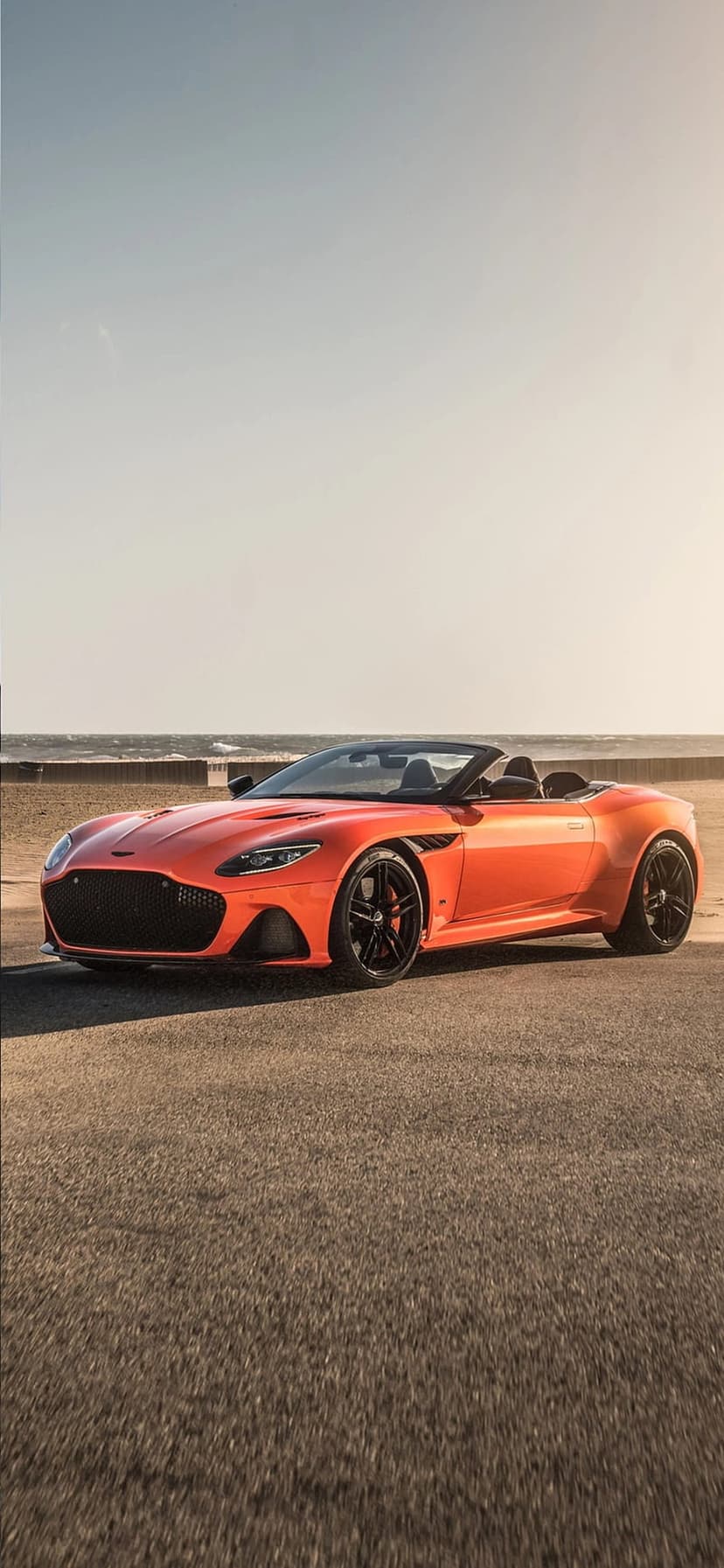 Orange Aston Martin convertible parked on a seaside road with black rims and a calm blue ocean in the background.