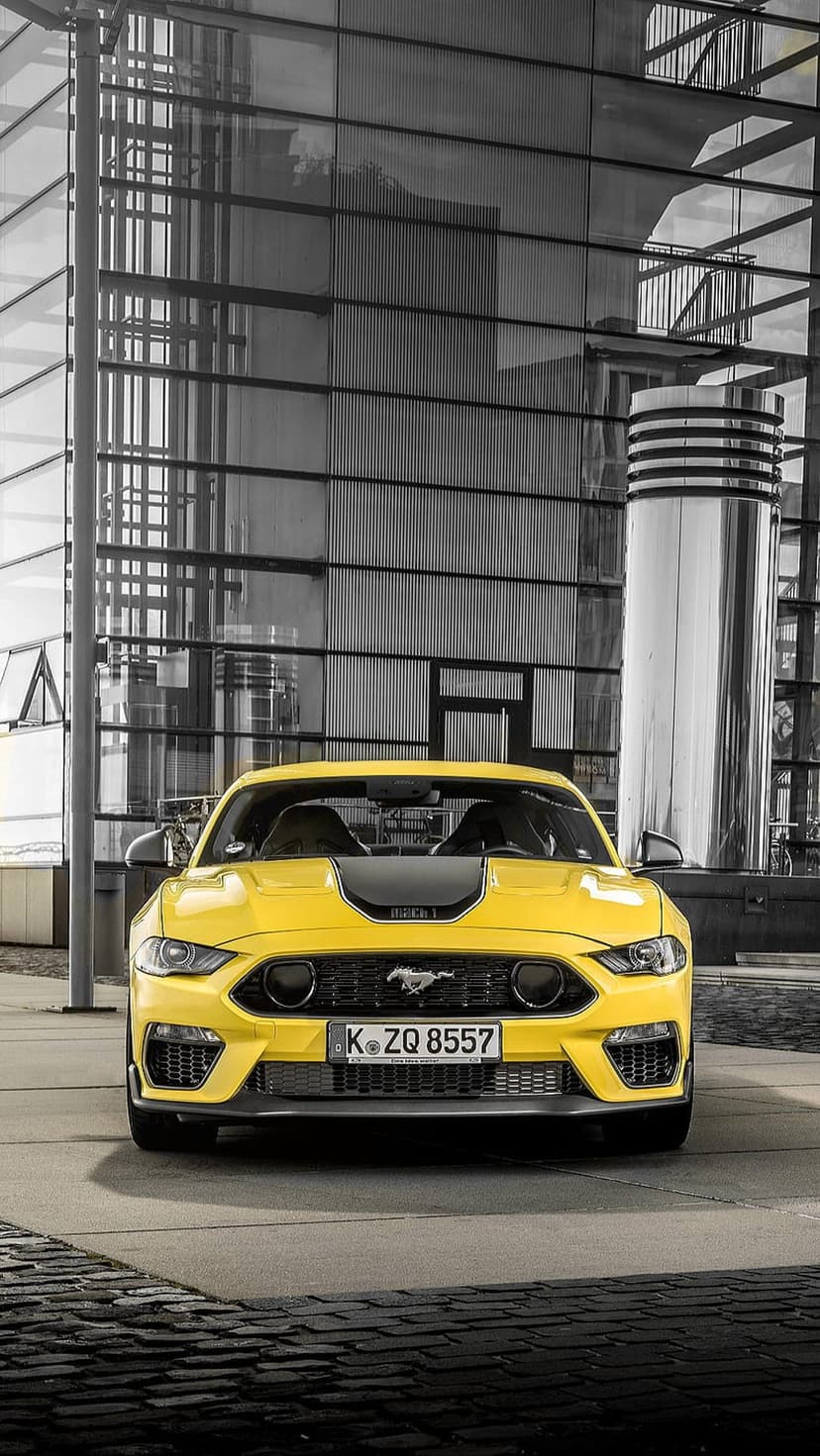 Yellow Ford Mustang Mach 1 sports car parked in front of a modern glass skyscraper with a grayscale finish.